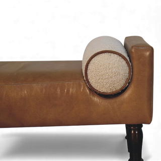 Side profile of a tan upholstered bench with cylindrical cream bouclé pillows and carved wooden legs.