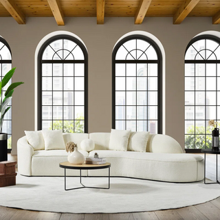White curved boucle sectional sofa styled in sophisticated living space with tall grid windows, round coffee table, and indoor plants, showcasing its flowing organic silhouette against neutral backdrop