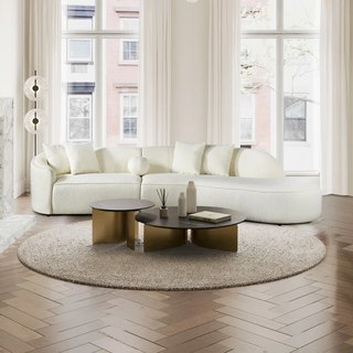 White boucle curved sectional styled in airy space with tall windows, herringbone wood floors, round area rug, and modern brass coffee tables creating elegant contemporary living arrangement